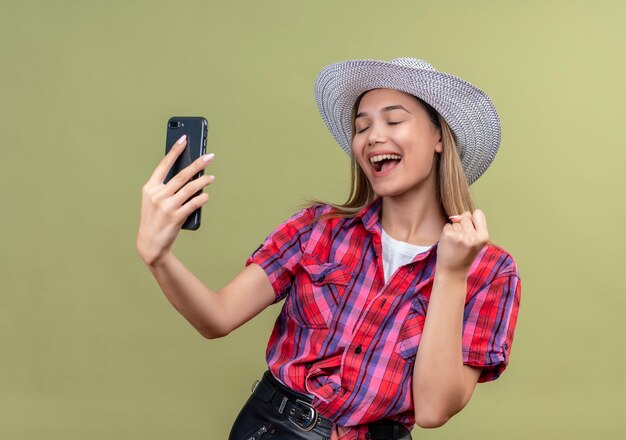 Une très heureuse belle jeune femme dans une chemise à carreaux à la recherche de téléphone mobile tout en levant le poing fermé