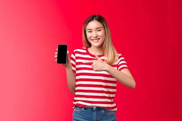 Très satisfaite joyeuse jolie fille blonde asiatique souriante montre largement l'affichage du smartphone pointant le téléphone...