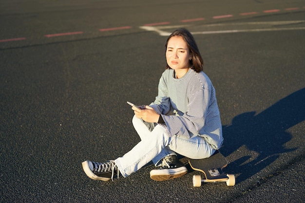 Triste adolescente asiatique assise sur une planche à roulettes a l'air bouleversée tient un téléphone portable et fronce les sourcils