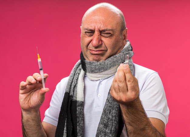 triste adulte malade caucasien avec un foulard autour du cou tenant une seringue et regardant une ampoule isolée sur un mur rose avec un espace de copie