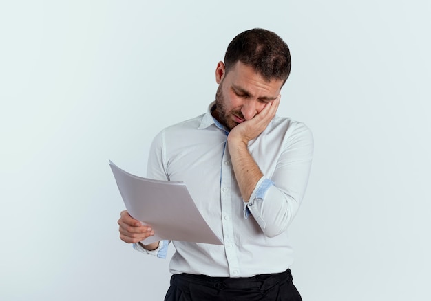 Triste bel homme met la main sur le visage tenant des feuilles de papier isolé sur mur blanc