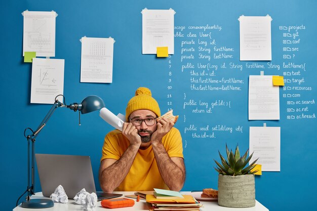 Triste étudiant frustré se prépare à l'examen en technologie informatique, tient du papier plié et mange un sandwich, les mains sous le menton et regarde malheureusement la caméra