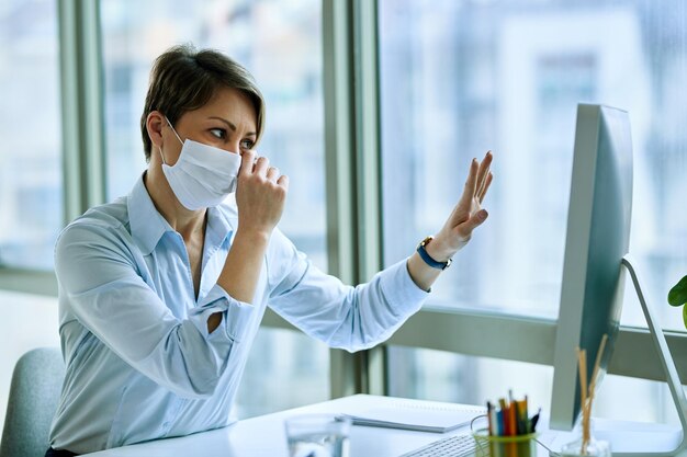 Triste femme d'affaires avec masque facial agitant tout en faisant un appel vidéo sur un ordinateur de bureau au bureau