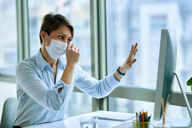 Triste femme d'affaires avec masque facial agitant tout en faisant un appel vidéo sur un ordinateur de bureau au bureau