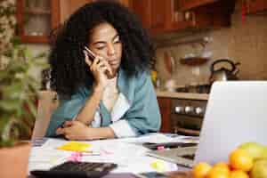 Photo gratuite triste femme africaine avec une coiffure afro assise dans la cuisine devant un ordinateur portable, parlant au téléphone portable de son mari, lui disant que leur famille sera bientôt expulsée en raison du non-paiement du loyer