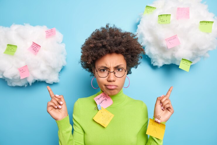 Photo gratuit triste femme afro-américaine frustrée point ci-dessus sur les nuages avec des notes autocollantes des tâches écrites à faire comme beaucoup de travail porte des lunettes rondes et un col roulé vert isolé sur un mur bleu