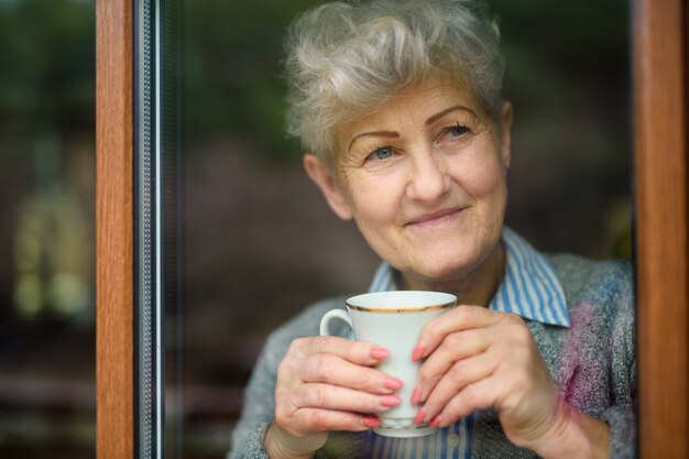 Triste femme âgée avec café regardant à l'intérieur à la maison, virus corona et concept de quarantaine.