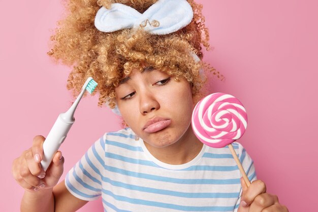 Triste femme frustrée aux cheveux bouclés regarde malheureusement la sucette veut manger sucré mais ne peut pas car elle a un effet nocif sur les dents tient une brosse à dents électrique habillée avec désinvolture isolée sur fond rose