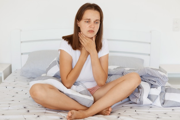 Triste femme malade et malheureuse aux cheveux noirs portant un t-shirt blanc décontracté assis sur le lit avec les jambes croisées, touchant son cou, souffrant de maux de gorge, fronçant les sourcils.