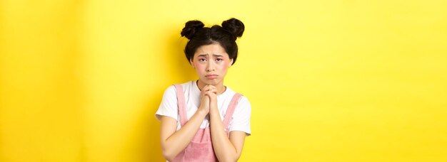 Photo gratuite triste fille asiatique implorant de l'aide vous suppliant de bouder et de pleurnicher debout sur fond jaune