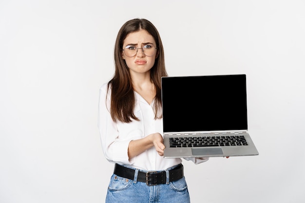 Triste fille brune, étudiante montrant un écran d'ordinateur portable et grimaçant contrarié, debout déçu sur fond blanc