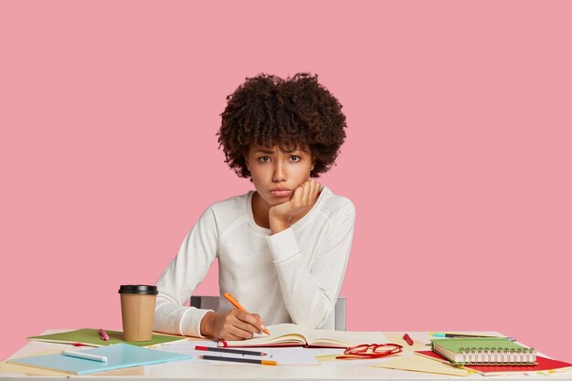 Triste fille étudiante stressante posant au bureau contre le mur rose