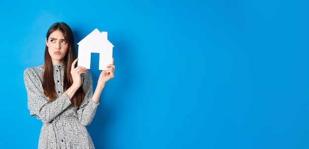 Photo gratuite triste fille de l'immobilier en fronçant les sourcils regardant de côté avec un visage injuste et bouleversé montrant la découpe de la maison en papier st