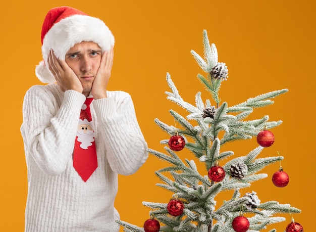 Triste jeune beau mec portant chapeau de Noël et cravate de père Noël debout près de sapin de Noël décoré en gardant les mains sur le visage en regardant la caméra isolée sur fond orange