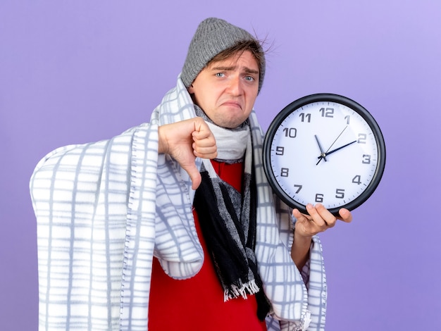 Triste jeune bel homme malade blonde portant un chapeau d'hiver et une écharpe enveloppée dans un plaid tenant horloge regardant la caméra montrant le pouce vers le bas isolé sur fond violet