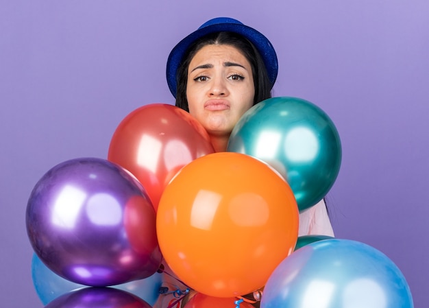 Triste jeune belle femme portant un chapeau de fête debout derrière des ballons isolés sur un mur bleu