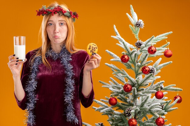 Triste jeune belle fille debout à proximité de l'arbre de Noël portant une robe rouge et une couronne avec guirlande sur le cou tenant un verre de lait avec des biscuits isolés sur fond orange