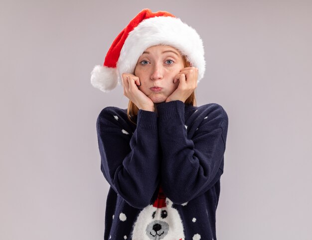 Triste jeune belle fille portant un chapeau de Noël et des lunettes mettant les mains sur les joues isolé sur fond blanc