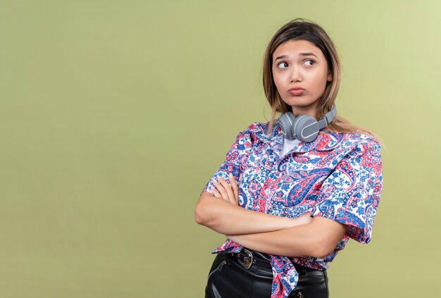 Une triste jeune femme en chemise imprimée paisley portant des écouteurs tenant les mains jointes sur un mur vert