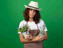 Photo gratuite triste jeune femme jardinière en uniforme portant chapeau de jardinage tenant et traversant des fleurs dans des pots de fleurs