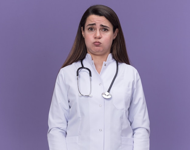 Triste jeune femme médecin portant une robe médicale avec un stéthoscope soufflant des joues isolées sur un mur violet avec espace de copie