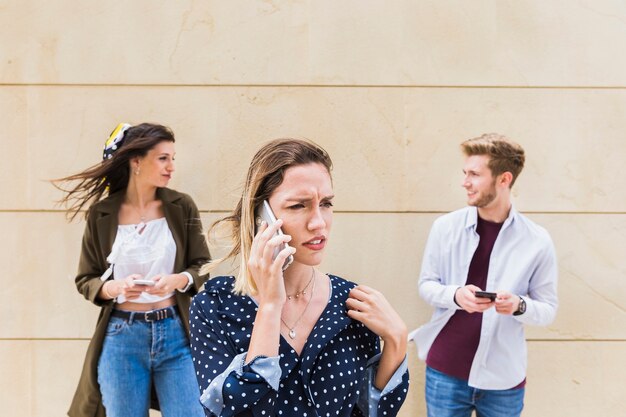 Triste jeune femme parlant sur téléphone portable debout devant des amis qui se regardent