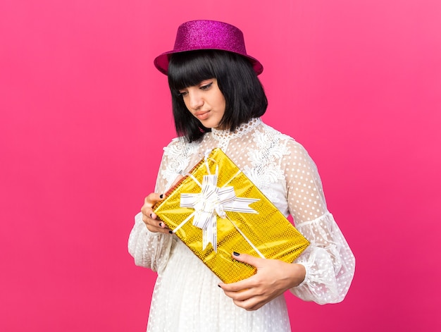 Photo gratuite triste jeune fêtarde portant un chapeau de fête tenant un paquet cadeau regardant vers le bas avec les lèvres pincées isolées sur un mur rose avec espace de copie