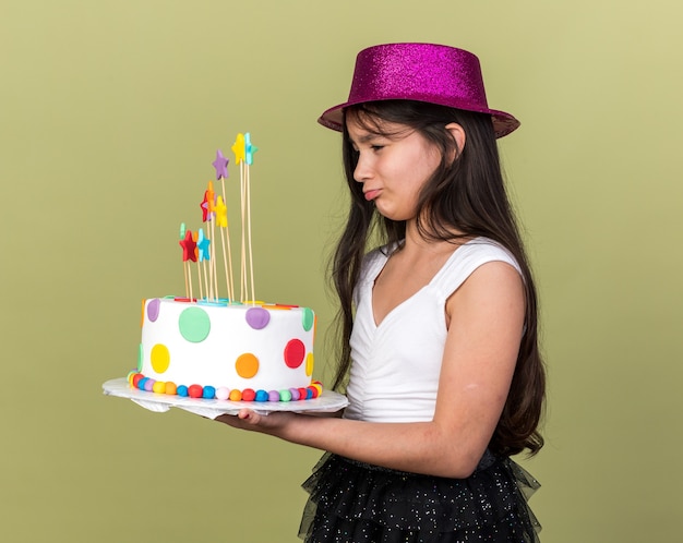 triste jeune fille caucasienne avec chapeau de fête violet tenant et regardant un gâteau d'anniversaire isolé sur un mur vert olive avec espace de copie