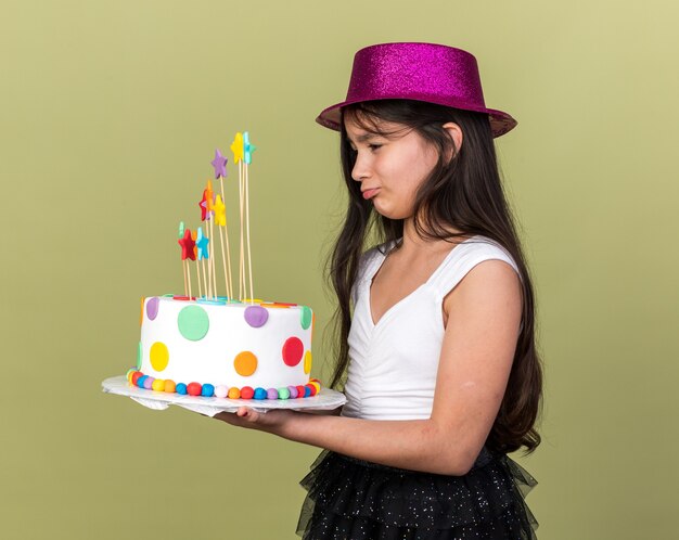 triste jeune fille caucasienne avec chapeau de fête violet tenant et regardant un gâteau d'anniversaire isolé sur un mur vert olive avec espace de copie