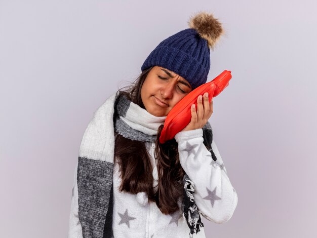 Triste jeune fille malade aux yeux fermés portant un chapeau d'hiver avec écharpe mettant le sac d'eau chaude sur la joue isolé sur fond blanc