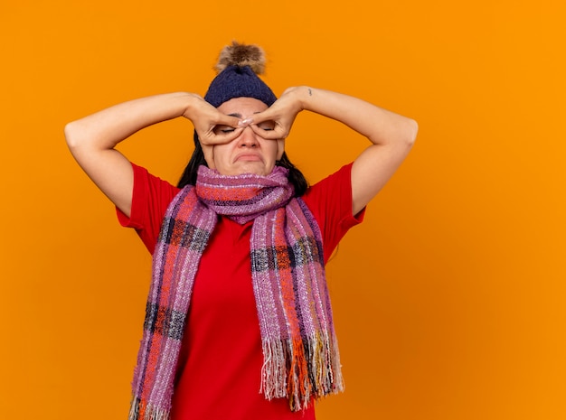 Photo gratuite triste jeune fille malade caucasienne portant chapeau d'hiver et écharpe faisant masque avec les mains avec les yeux fermés isolé sur fond orange avec espace copie