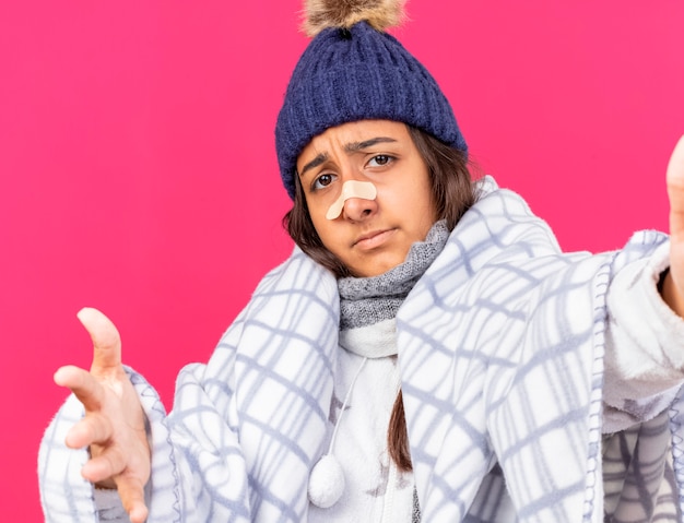 Photo gratuite triste jeune fille malade portant chapeau d'hiver avec écharpe enveloppée de plaid avec du plâtre sur le nez tenant la caméra isolée sur fond rose