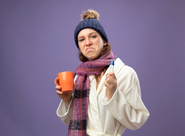 Triste jeune fille malade portant une robe blanche et un chapeau d'hiver avec écharpe tenant une tasse de thé avec thermomètre isolé sur violet