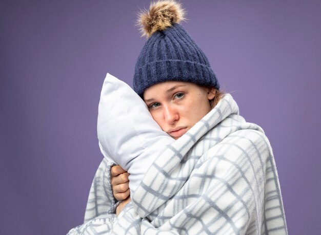 Triste jeune fille malade portant une robe blanche et un chapeau d'hiver avec un foulard enveloppé dans un oreiller étreint à carreaux isolé sur violet