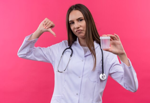 Triste jeune fille médecin portant stéthoscope robe médicale tenant vide peut son pouce vers le bas sur fond rose isolé