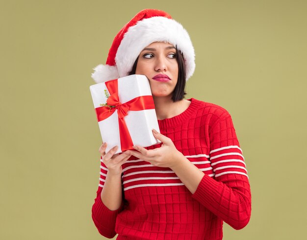 Triste jeune fille portant bonnet de Noel tenant un paquet cadeau touchant le visage avec elle à côté isolé sur fond vert olive avec espace copie