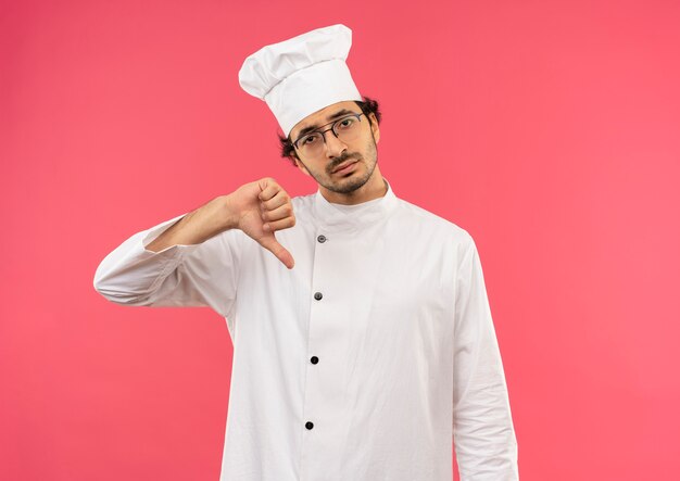 Triste jeune homme cuisinier portant l'uniforme de chef et des lunettes son pouce vers le bas isolé sur un mur rose