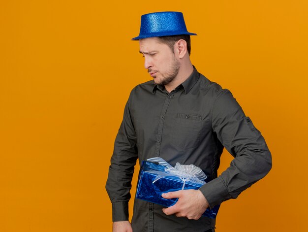 Triste jeune homme de fête portant un chapeau bleu tenant une boîte-cadeau isolée sur orange