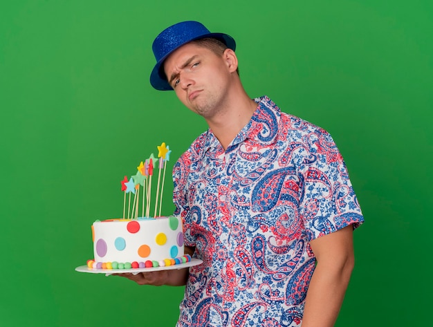 Triste jeune homme de fête portant un chapeau bleu tenant un gâteau isolé sur vert