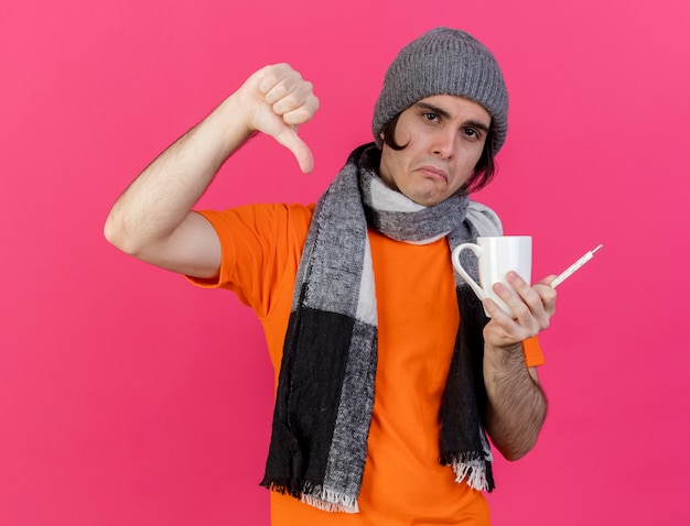 Triste jeune homme malade portant chapeau d'hiver avec écharpe tenant une tasse de thé avec thermomètre montrant le pouce vers le bas isolé sur fond rose