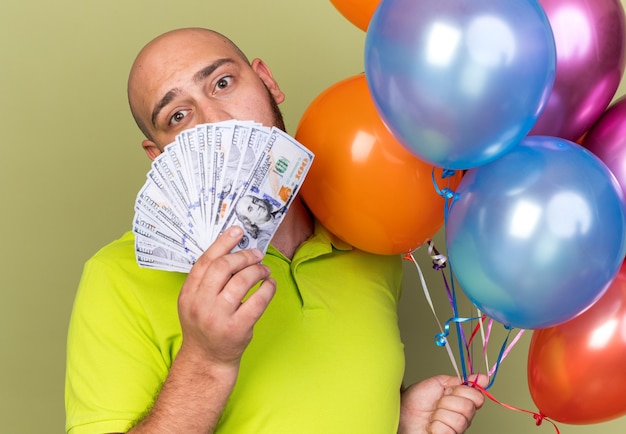 Triste jeune homme portant un t-shirt jaune tenant des ballons le visage couvert d'argent isolé sur un mur vert olive