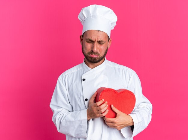 Triste jeune homme de race blanche cuisinier en uniforme de chef et casquette tenant en forme de coeur pleurant les yeux fermés isolés sur le mur rose