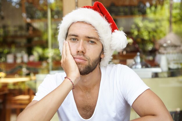 Triste jeune homme de race blanche portant un t-shirt décontracté et un chapeau de père Noël rouge avec une fourrure blanche faisant la moue, l'air fatigué et ennuyé, se sentant seul