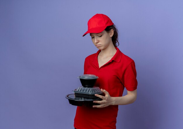 Triste jeune jolie livreuse portant un uniforme rouge et une casquette tenant et regardant des contenants de nourriture isolés sur fond violet avec espace de copie