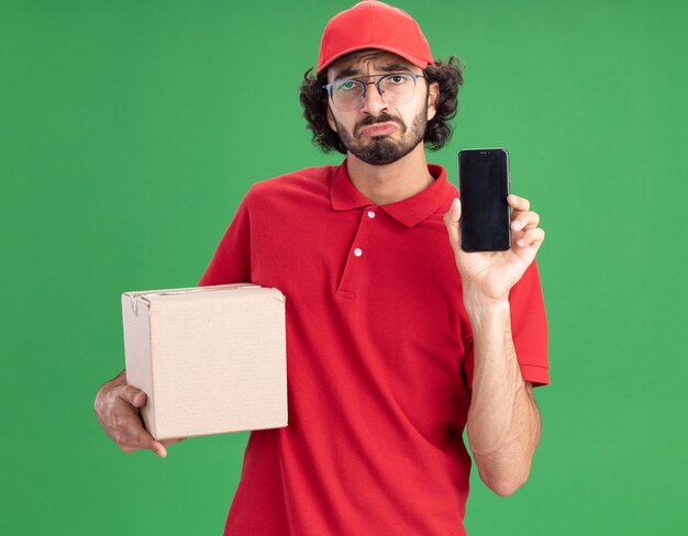 Triste jeune livreur caucasien en uniforme rouge et casquette portant des lunettes tenant une boîte à cartes montrant un téléphone portable isolé sur un mur vert