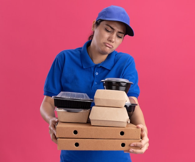 Triste jeune livreuse en uniforme avec casquette tenant des contenants de nourriture sur des boîtes à pizza isolées sur un mur rose