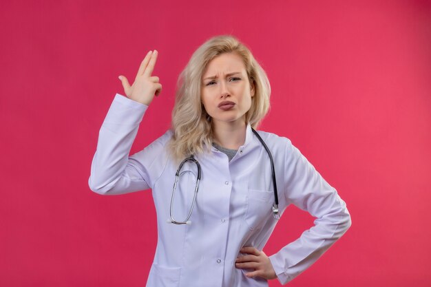 Triste jeune médecin portant un stéthoscope en robe médicale montrant le geste du pistolet sur fond rouge