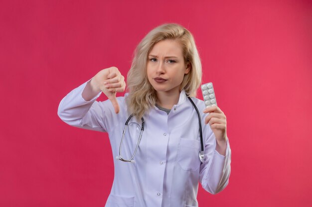 Triste jeune médecin portant un stéthoscope en robe médicale tenant des pilules son pouce vers le bas sur fond rouge