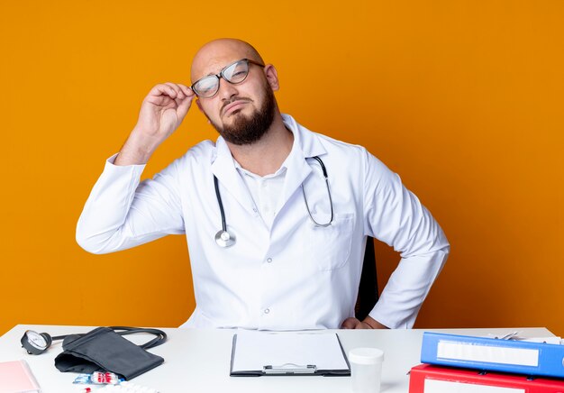 Triste jeune médecin de sexe masculin chauve portant robe médicale et stéthoscope assis au bureau avec des outils médicaux prendre des lunettes sur orange