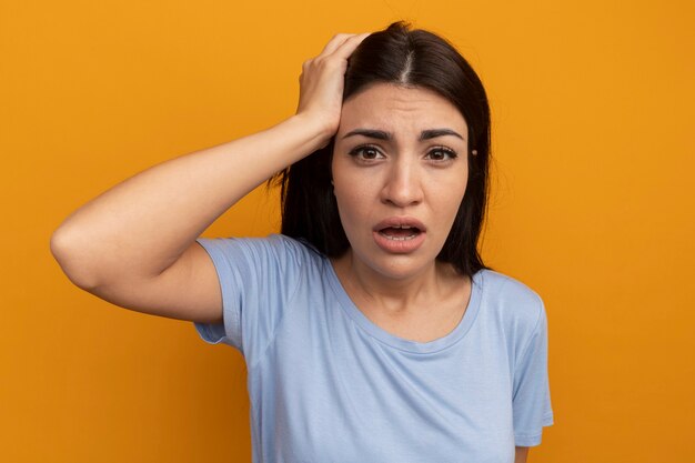 Triste jolie femme brune met la main sur la tête et regarde à l'avant isolé sur mur orange